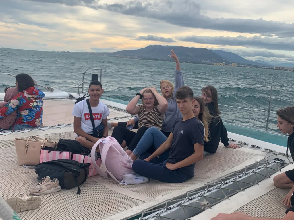 Jugendliche sitzen auf dem Vordeck eines Segelboots, dahinter die Küste.